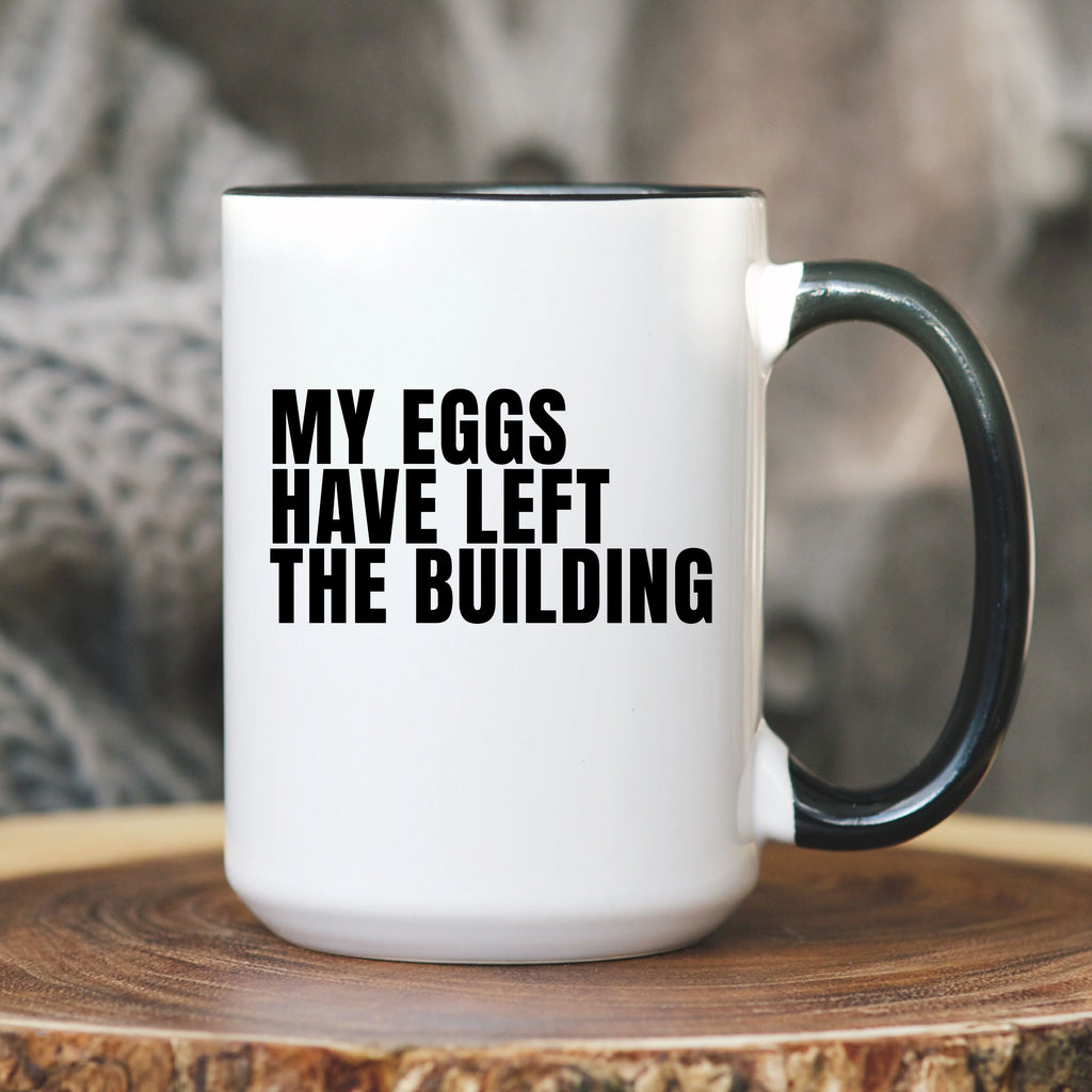 White mug with black text 'MY EGGS HAVE LEFT THE BUILDING' on a wooden surface.