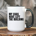White mug with black text 'MY EGGS HAVE LEFT THE BUILDING' on a wooden surface.